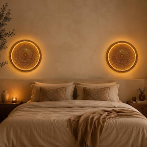 May include: Two circular wall-mounted light fixtures with intricate perforated patterns emit a warm, golden glow. The lights are positioned above a bed featuring a neutral-toned duvet, two matching pillows, and a draped throw blanket.