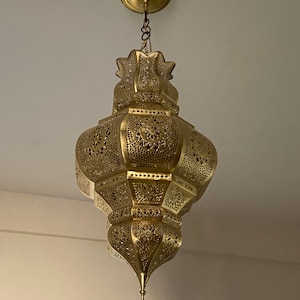May include: Ornate gold-colored pendant light fixture. The lamp has a detailed, openwork design with floral patterns. It is suspended from the ceiling by a chain and a circular canopy. The light fixture is a decorative element for interior spaces.