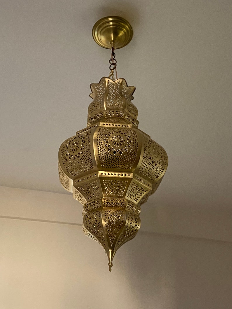 May include: Ornate gold-colored pendant light fixture. The lamp has a detailed, openwork design with floral patterns. It is suspended from the ceiling by a chain and a circular canopy. The light fixture is a decorative element for interior spaces.