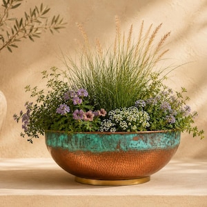 May include: A round, hammered copper planter bowl featuring a vibrant turquoise patina finish along the upper rim. The planter is filled with a variety of green grasses, small purple flowers, and white blossoms, resting on a circular gold base.
