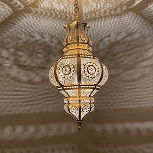 May include: Ornate, gold-colored pendant light with intricate cut-out patterns. The lamp is suspended from the ceiling by a chain and emits a warm glow, casting decorative shadows on the surrounding surface. The design features floral and geometric shapes.