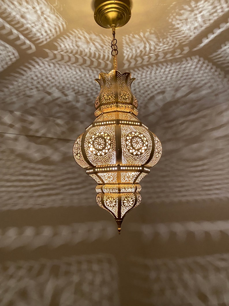 May include: Ornate, gold-colored pendant light with intricate cut-out patterns. The lamp is suspended from the ceiling by a chain and emits a warm glow, casting decorative shadows on the surrounding surface. The design features floral and geometric shapes.