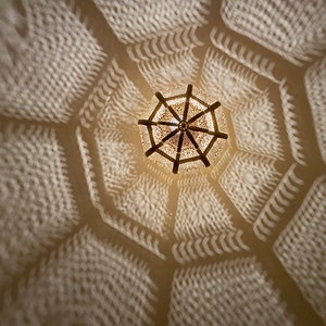 May include: An overhead shot of a decorative, ornate light fixture casting intricate geometric shadows on a white ceiling. The light fixture is a warm, golden color, and the shadows create a repeating pattern.