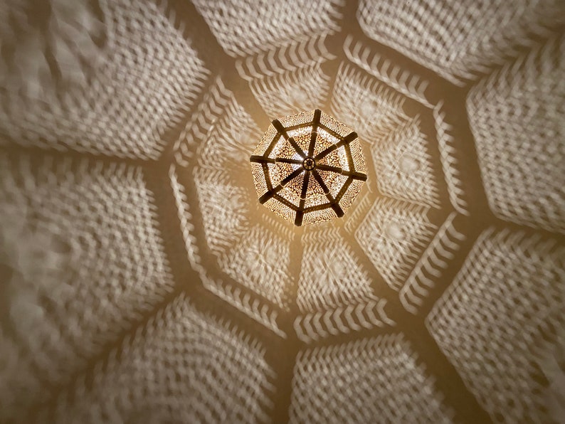 May include: An overhead shot of a decorative, ornate light fixture casting intricate geometric shadows on a white ceiling. The light fixture is a warm, golden color, and the shadows create a repeating pattern.