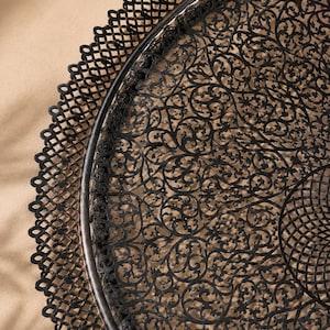 May include: A round black metal tray featuring an intricate, laser-cut filigree pattern across its surface. The tray has a raised, scalloped edge with a repeating lattice design that frames the detailed, swirling floral-inspired metalwork.