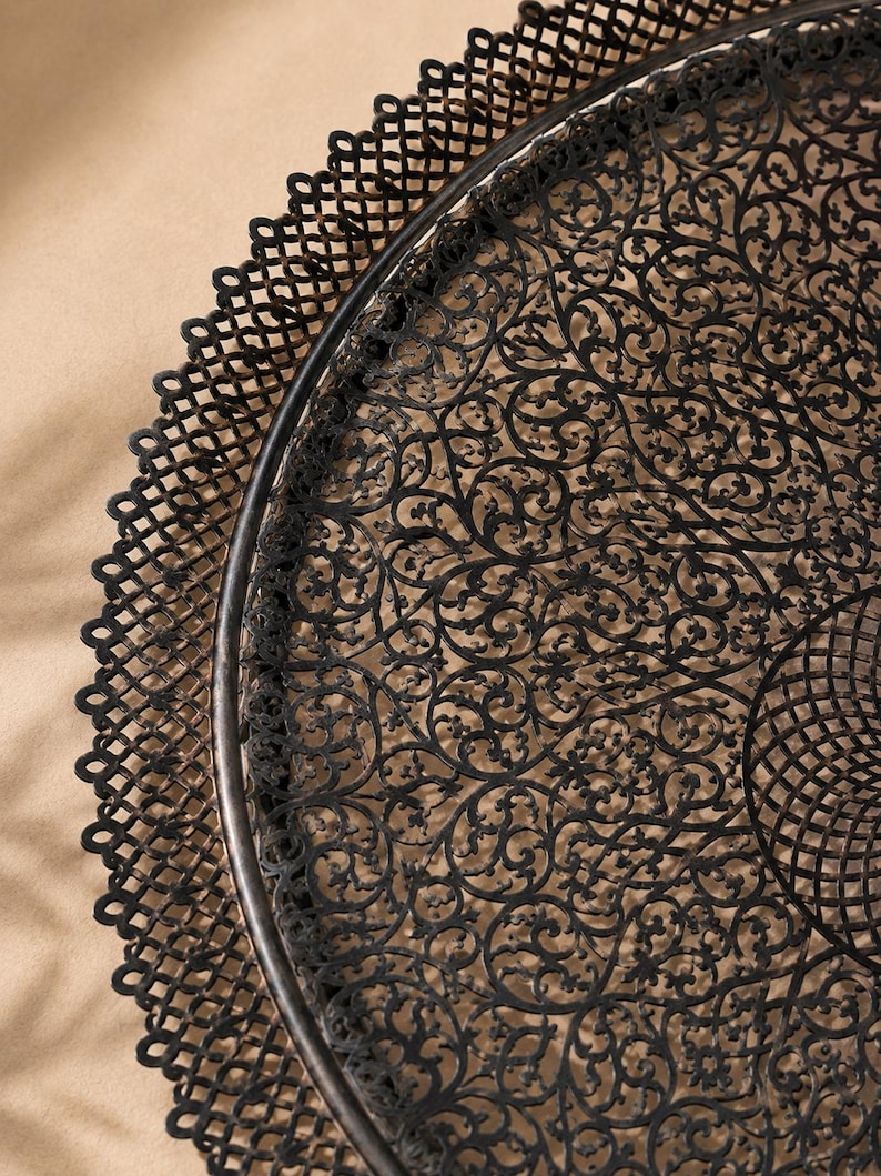 May include: A round black metal tray featuring an intricate, laser-cut filigree pattern across its surface. The tray has a raised, scalloped edge with a repeating lattice design that frames the detailed, swirling floral-inspired metalwork.