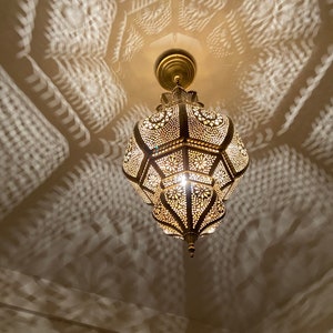 May include: Ornate, gold-colored pendant light fixture with intricate cut-out patterns. The light casts decorative shadows on the ceiling and walls. The fixture is suspended from a matching gold ceiling mount.