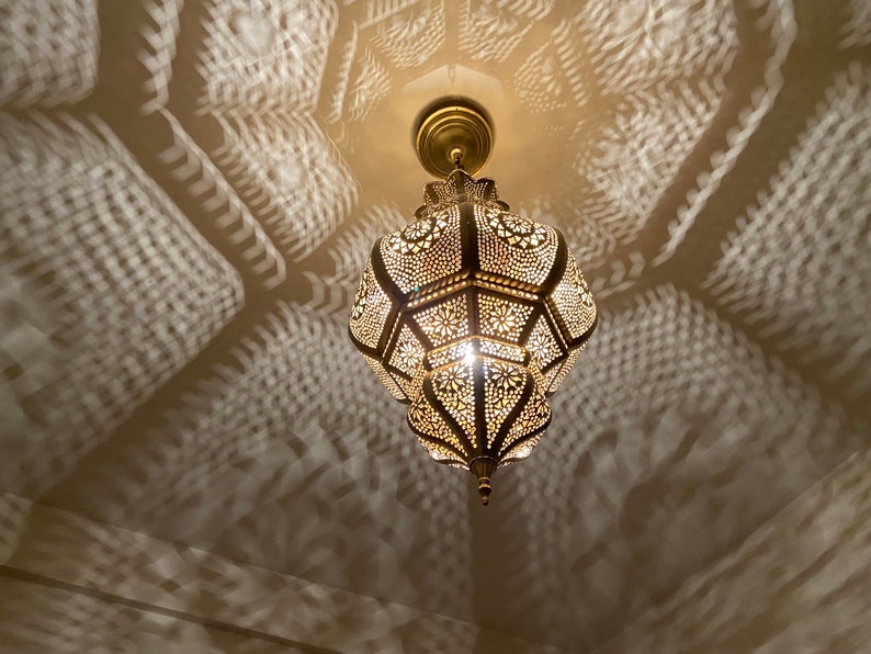 May include: Ornate, gold-colored pendant light fixture with intricate cut-out patterns. The light casts decorative shadows on the ceiling and walls. The fixture is suspended from a matching gold ceiling mount.