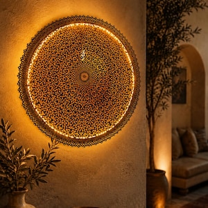 May include: A circular wall-mounted light fixture featuring an intricate, dark-colored metal cutout pattern with a fine, lace-like design. The fixture emits a warm, golden glow from behind its perimeter, casting light onto the wall surface.