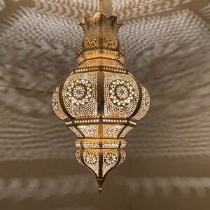 May include: Ornate, gold-colored pendant lamp with intricate cut-out patterns. The lamp is suspended from the ceiling, casting decorative shadows on the wall. The design features floral and geometric shapes, creating a warm, inviting ambiance.