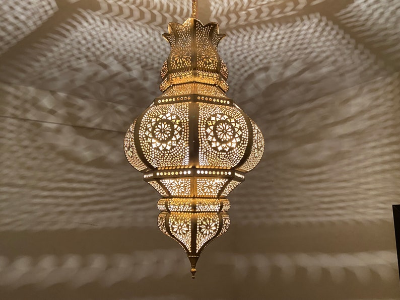 May include: Ornate, gold-colored pendant lamp with intricate cut-out patterns. The lamp is suspended from the ceiling, casting decorative shadows on the wall. The design features floral and geometric shapes, creating a warm, inviting ambiance.