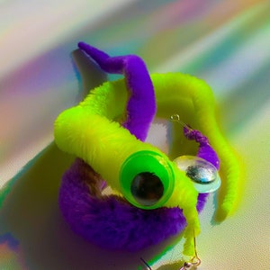 May include: A pair of fuzzy worm earrings with googly eyes. One worm is purple and the other is neon green.