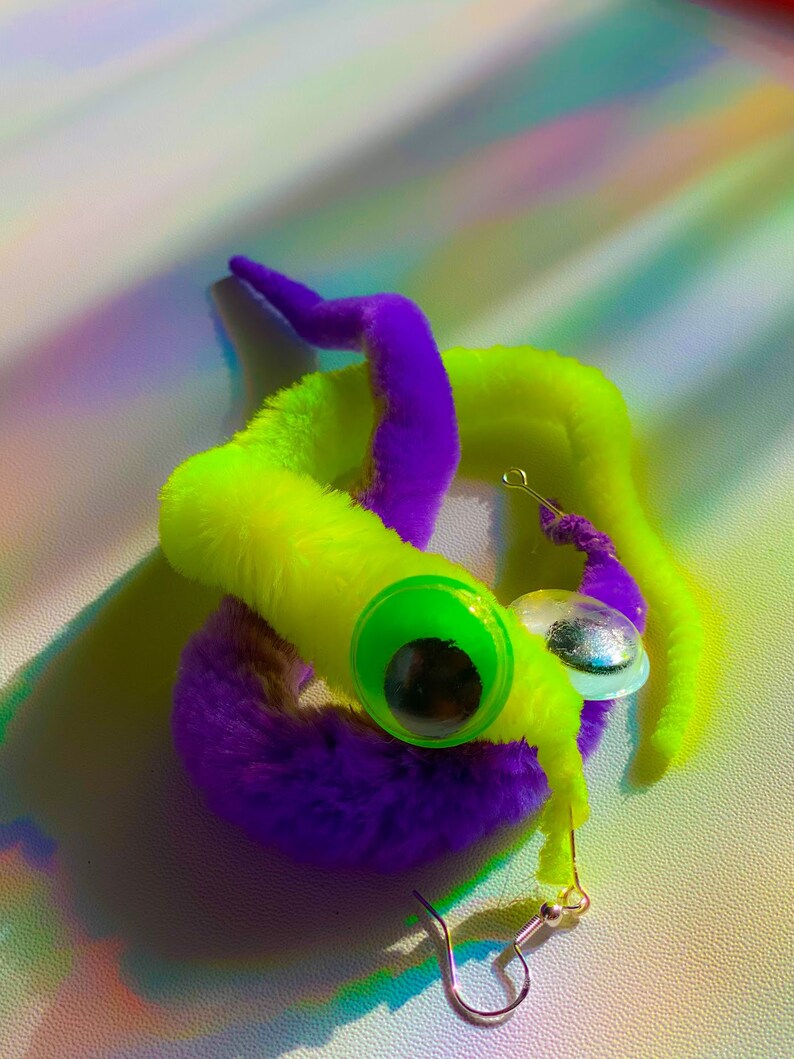 May include: A pair of fuzzy worm earrings with googly eyes. One worm is purple and the other is neon green.