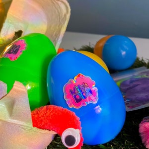 May include: A blue plastic egg with a holographic sticker that says "Miss & Dazzle" on it. The egg is sitting on a bed of green grass. There is a red fuzzy worm with a black eye poking out of a broken egg shell.