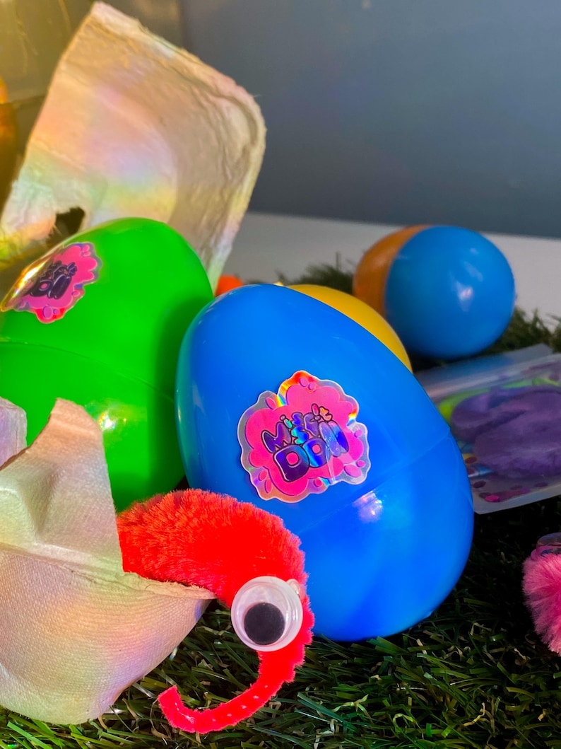 May include: A blue plastic egg with a holographic sticker that says "Miss & Dazzle" on it. The egg is sitting on a bed of green grass. There is a red fuzzy worm with a black eye poking out of a broken egg shell.