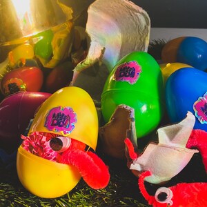 May include: A yellow plastic egg with a red fuzzy worm inside. The egg is open and has a sticker that says "Mini Boi" on it. There are other plastic eggs in the background, some are green, blue, and purple.