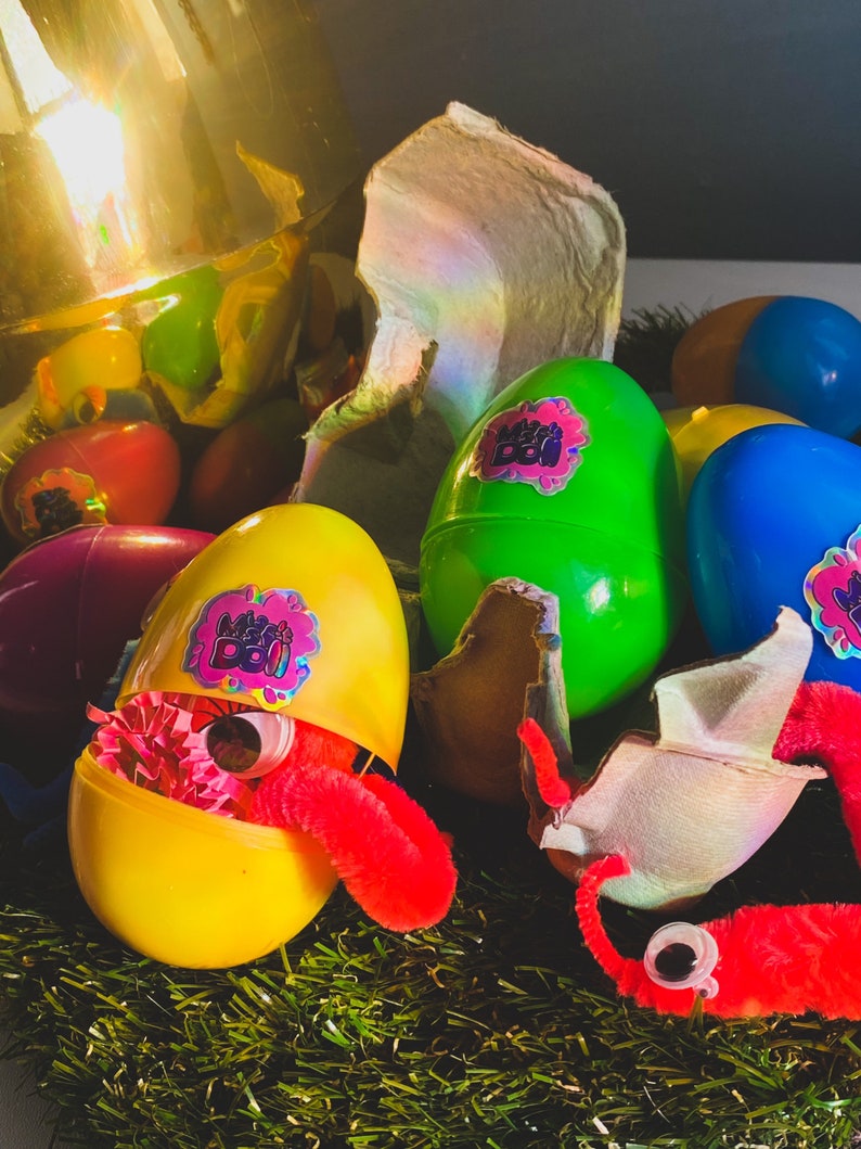 May include: A yellow plastic egg with a red fuzzy worm inside. The egg is open and has a sticker that says "Mini Boi" on it. There are other plastic eggs in the background, some are green, blue, and purple.