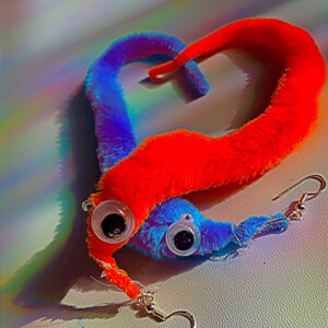 May include: A pair of fuzzy worm earrings with googly eyes. The worm on the left is red and the worm on the right is blue. The earrings are hanging from silver hooks.