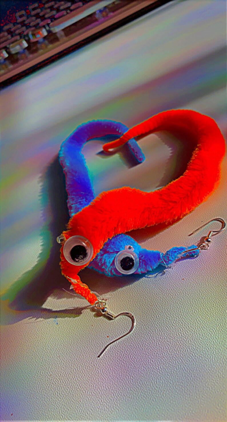 May include: A pair of fuzzy worm earrings with googly eyes. The worm on the left is red and the worm on the right is blue. The earrings are hanging from silver hooks.