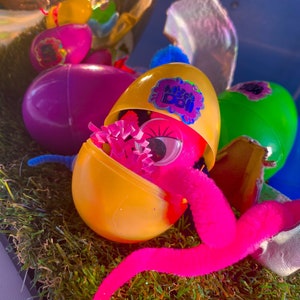 May include: A yellow plastic egg with a pink fuzzy worm coming out of it. The egg is open and has a pink eye and pink and purple tissue paper inside. The egg has a sticker that says "Mystery Boo!" on it.