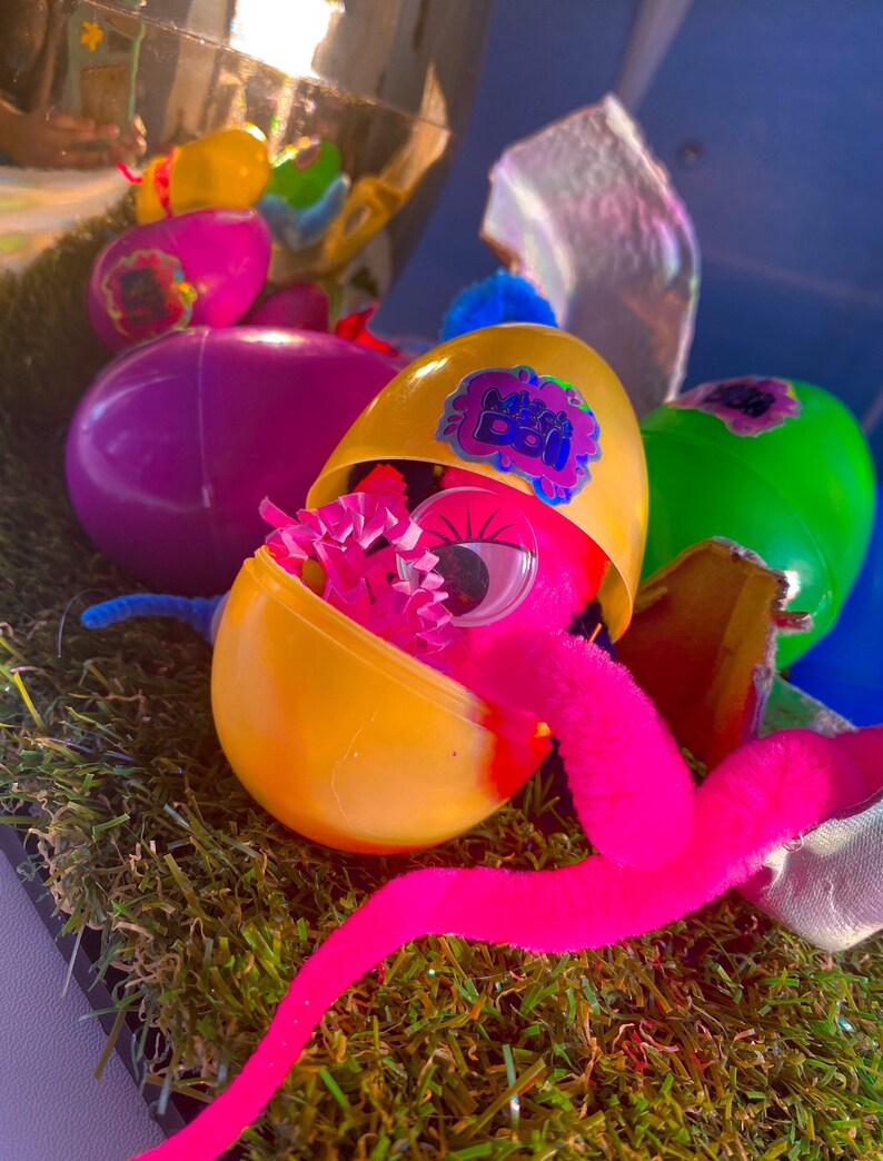 May include: A yellow plastic egg with a pink fuzzy worm coming out of it. The egg is open and has a pink eye and pink and purple tissue paper inside. The egg has a sticker that says "Mystery Boo!" on it.