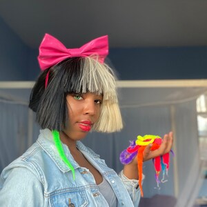 May include: A person with black and white hair wearing a pink bow headband and a denim jacket. They are holding a bunch of colorful fuzzy worm toys in their hand.