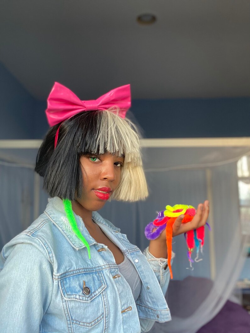 May include: A person with black and white hair wearing a pink bow headband and a denim jacket. They are holding a bunch of colorful fuzzy worm toys in their hand.