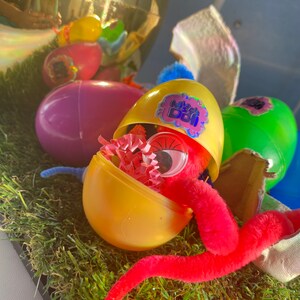 May include: A yellow plastic egg with a pink and purple sticker that says "My Bestie" is open and reveals a red fuzzy worm and pink and white tissue paper inside.