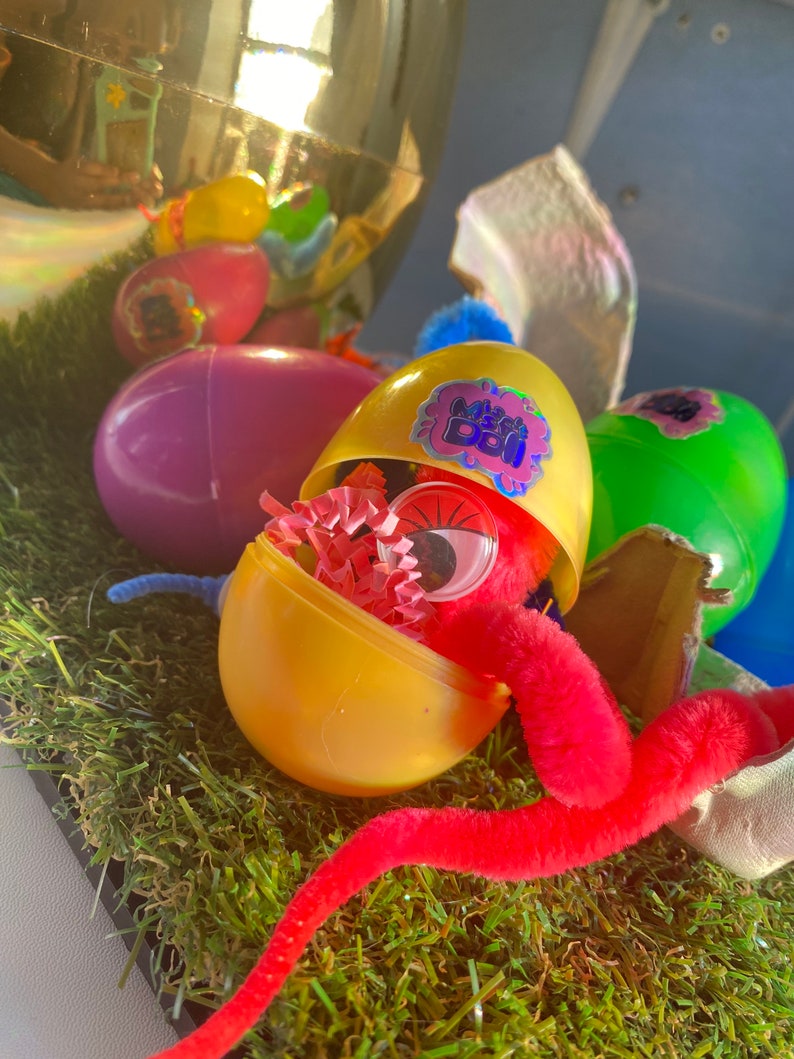 May include: A yellow plastic egg with a pink and purple sticker that says "My Bestie" is open and reveals a red fuzzy worm and pink and white tissue paper inside.