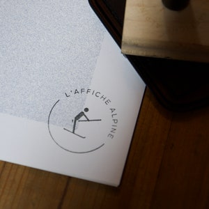 Puede incluir: Un p&oacute;ster blanco con un logotipo en blanco y negro que dice "L'AFFICHE ALPINE" en un c&iacute;rculo. El logotipo presenta a un esquiador en una silueta negra.