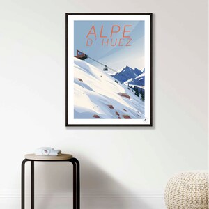 Puede incluir: Un p&oacute;ster enmarcado en negro con una ilustraci&oacute;n azul y blanca de un telesilla subiendo una monta&ntilde;a nevada. El texto "ALPE D'HUEZ" est&aacute; en letras rojas en la parte superior del p&oacute;ster.