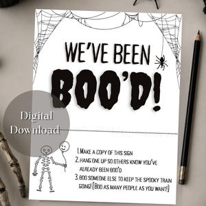 以下が含まれることがあります： WE'VE BEEN BOO'D!と書かれた黒い文字とハロウィーンゲームの説明が記載された白い長方形の看板。デザインには、四隅の黒いクモの巣のイラスト、小さなクモ、風船を持った骸骨が描かれています。