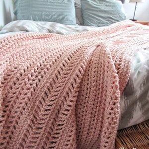 May include: A pink crocheted throw blanket with a chevron pattern is draped over a bed with gray and white bedding. The blanket is made with a soft yarn and has a delicate texture. The text "This Pixie Creates" is visible in the bottom right corner of the image.