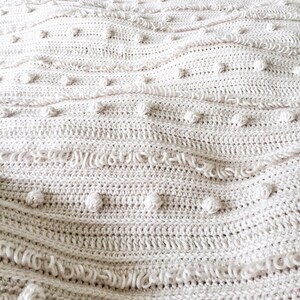 May include: A close-up of a cream-coloured crocheted throw blanket with a textured pattern of small bumps and loops.