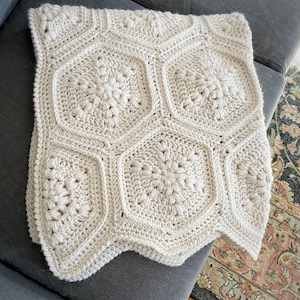 May include: A crocheted blanket with a geometric pattern in a cream color. The blanket is folded and placed on a gray couch.