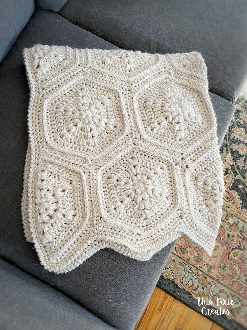 May include: A crocheted blanket with a geometric pattern in a cream color. The blanket is folded and placed on a gray couch.