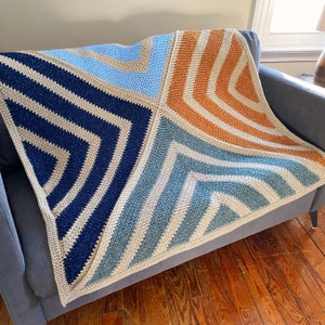May include: A crocheted throw blanket with a geometric pattern in shades of blue, beige, and orange. The blanket is folded and draped over a sofa.