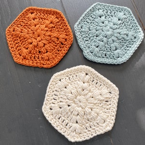 May include: Three crocheted hexagon coasters in different colors: orange, light blue, and cream.