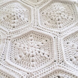 May include: A close-up of a crocheted blanket with a repeating pattern of white hexagonal shapes. Each hexagon features a raised, flower-like design in the center.