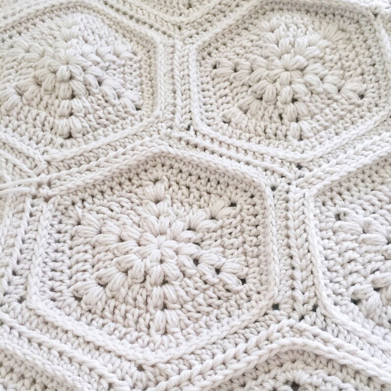 May include: A close-up of a crocheted blanket with a repeating pattern of white hexagonal shapes. Each hexagon features a raised, flower-like design in the center.