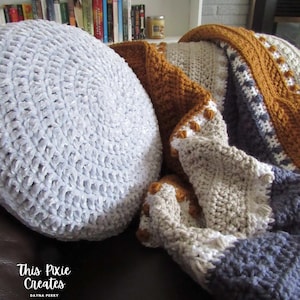 May include: A close-up of a crocheted throw blanket with a white, gray, and brown color scheme. The blanket is folded and draped over a light gray round pillow. The pillow is made of a soft, plush fabric. The text "This Pixie Creates" is visible in the lower left corner of the image.