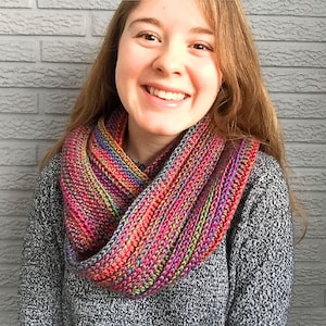 May include: A woman wearing a colorful crocheted infinity scarf. The scarf is made with a variety of shades of pink, red, orange, green, and purple yarn.