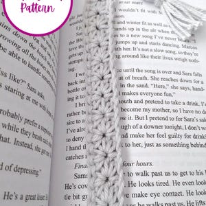 May include: A close-up of a white crochet bookmark with a tassel. The bookmark is made with a simple stitch pattern and has a delicate, feminine look. The bookmark is placed on a book with the text "Heart Bones" visible.