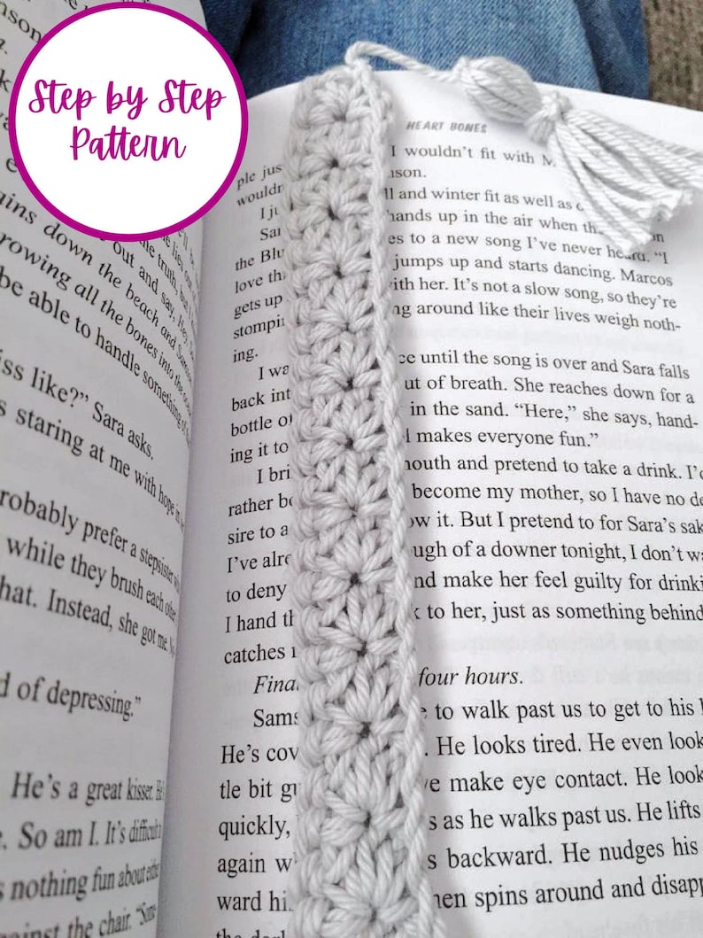 May include: A close-up of a white crochet bookmark with a tassel. The bookmark is made with a simple stitch pattern and has a delicate, feminine look. The bookmark is placed on a book with the text "Heart Bones" visible.
