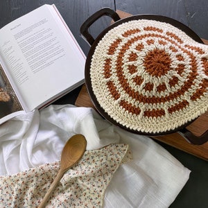 Puede incluir: Un posavasos de ganchillo en un esquema de color crema y marrón con un diseño circular. El posavasos está sobre una superficie de madera marrón oscuro. También hay un paño blanco y una cuchara de madera en la imagen.