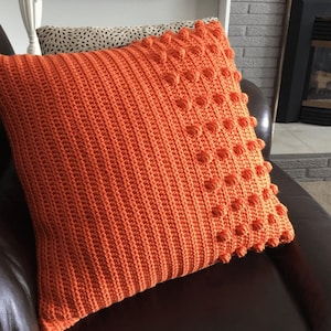 Puede incluir: Cojín de punto de ganchillo naranja con un diseño texturizado. El cojín tiene una textura en relieve, abultada en un lado y una textura lisa y acanalada en el otro.