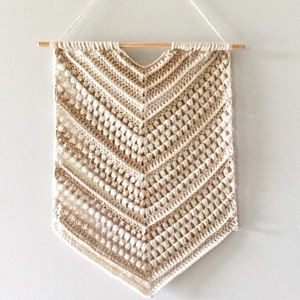 May include: A beige crocheted wall hanging with a chevron pattern and a wooden dowel at the top. The hanging has a textured, bumpy surface.