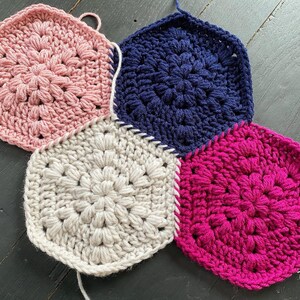May include: Four crocheted hexagons in pink, white, navy blue, and fuchsia, joined together to form a larger piece. The hexagons have a raised, textured pattern.