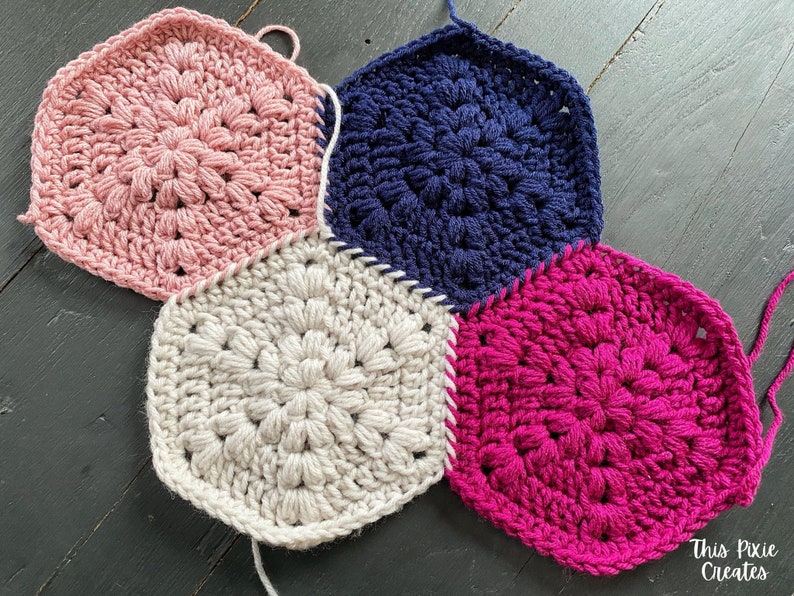 May include: Four crocheted hexagons in pink, white, navy blue, and fuchsia, joined together to form a larger piece. The hexagons have a raised, textured pattern.