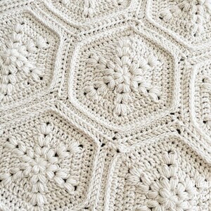 May include: A close-up of a crocheted blanket with a geometric pattern of beige hexagons. The blanket is made with a soft, textured yarn and has a subtle sheen.